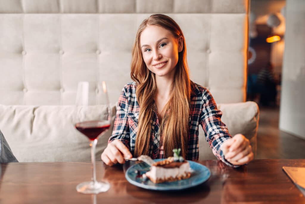Portrait Of Young Woman In Cafe, Sweet Cake And Red Wine In A Glass On The Table. Girl With Chocolate Dessert In Restaurant